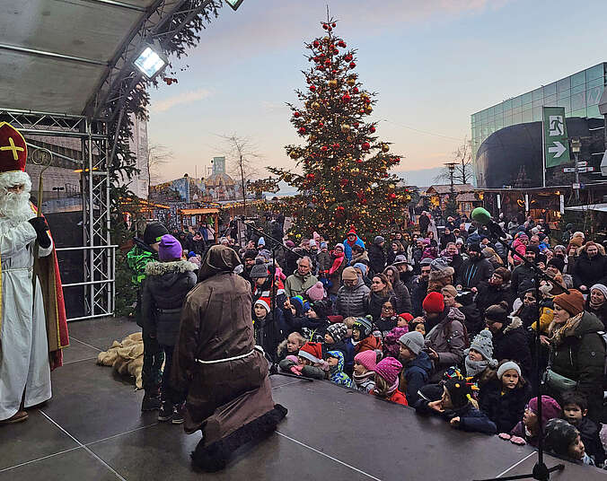 Der Nikolaus hält ein Buch in der Hand und viele Kinder stehen dichtgedrängt vor der Bühne