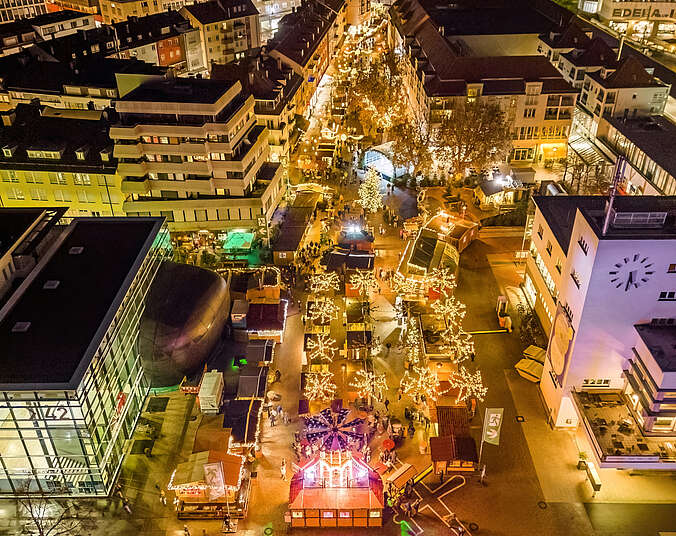 Blick auf den Weihnachtsmarkt mit vielen Lichtern. Rechts und links Häuser.